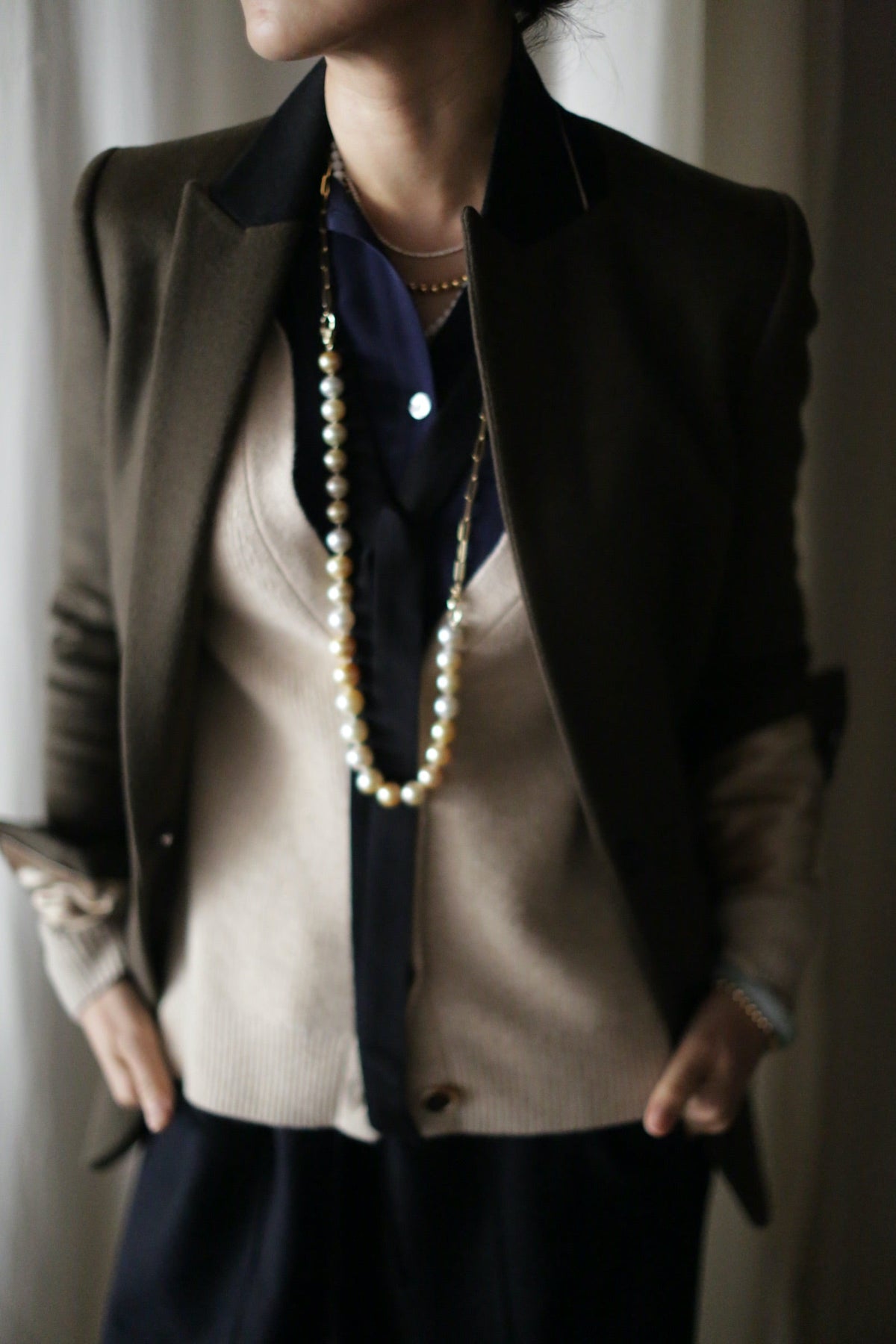 The Sloane | Olive / Black tuxedo jacket is worn over a beige, woven cashmere cardigan and a dark collared shirt, accessorized with a long pearl necklace.