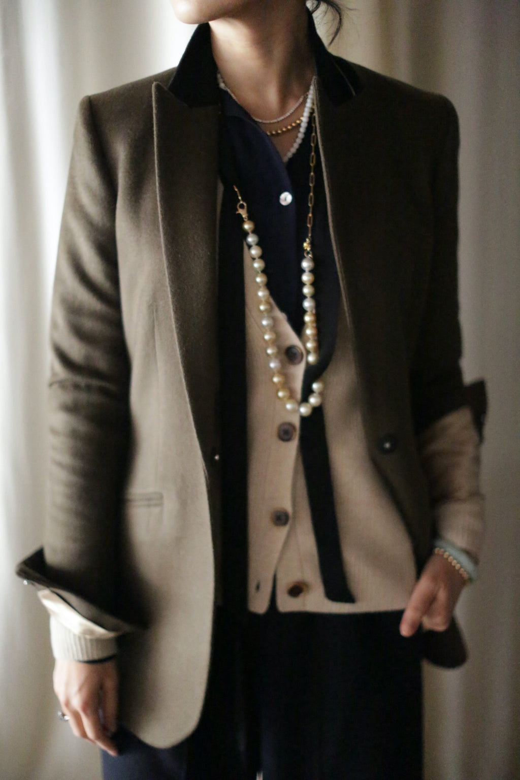 A person wears a Sloane | Taupe / Black equestrian length tuxedo jacket with a black collar over a beige cardigan, dark blue shirt, and a long pearl necklace.