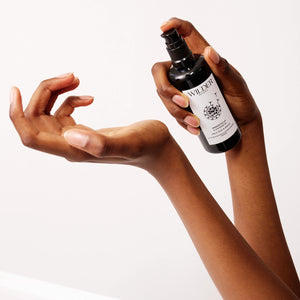 A close-up of a person spraying the Immortelle Cleansing Oil, packaged in a sleek black bottle labeled "Wilder," onto their hand. Infused with skin-renewing botanicals such as Helichrysum oil, this spray promises rejuvenation. The background is plain white, and the hands are positioned elegantly, one holding the bottle and the other poised to receive the luxurious cleansing oil.