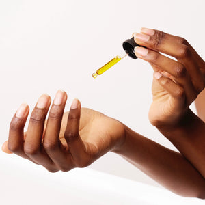 A dropper bottle is held by a person, dispensing a golden yellow liquid into their cupped palm. The hands are gracefully positioned against a minimalist, light-toned background, showcasing the Recovery Night Oil rich in phytonutrients.