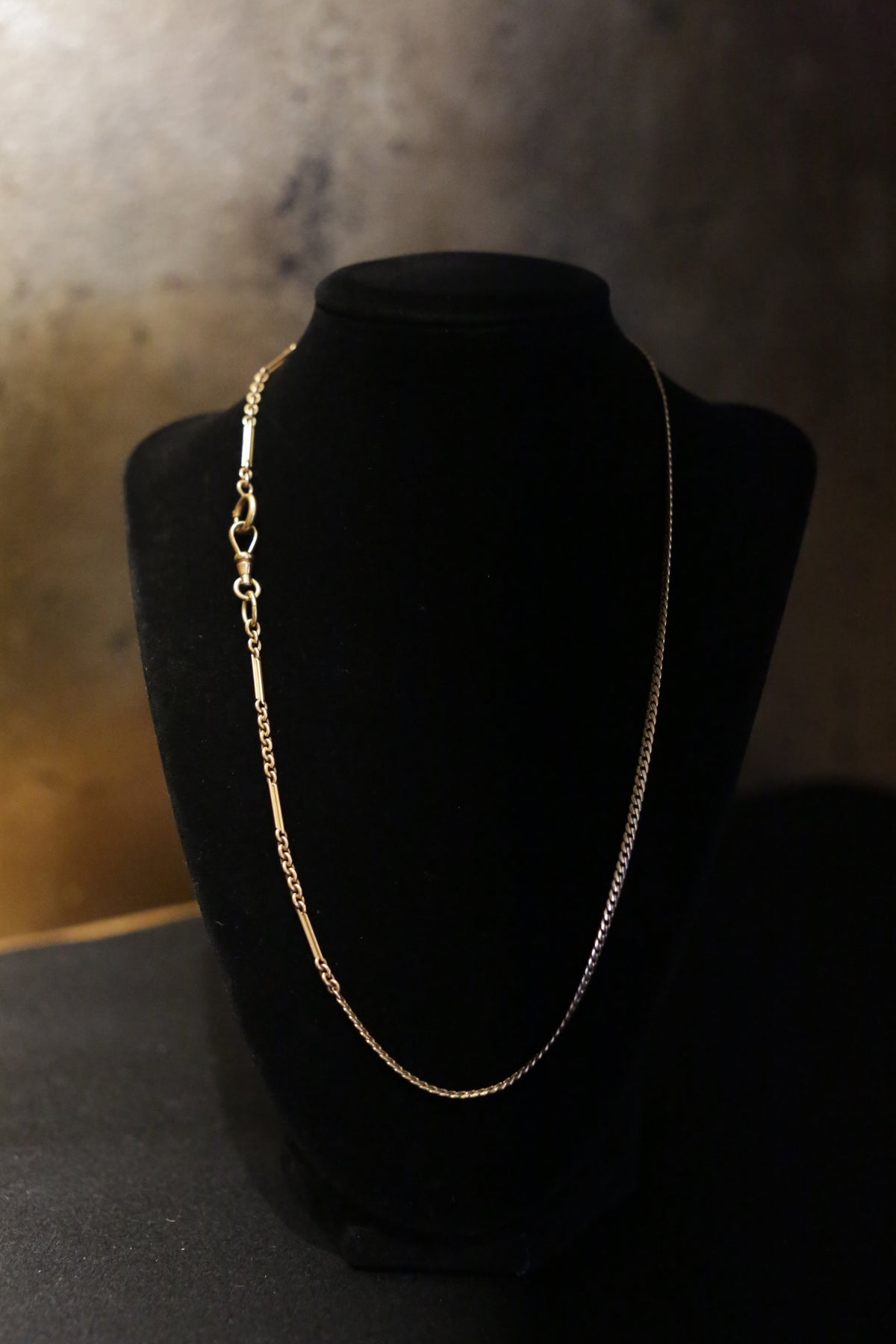 The Antique 14K Gold Bar and Link Chain c.1900 with a 12K Flat Curb Chain c.1880 is showcased on a black velvet stand, set against a darkly shaded background. This contrast enhances the intricate design and glistening texture of the antique jewelry pieces, highlighting their unique link styles and timeless elegance.
