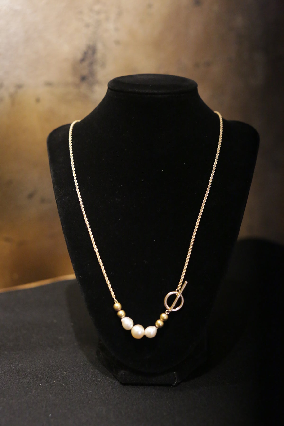 The Trio of Vintage Pearls and 14K Beads on a 14K Gold Chain with a toggle closure is elegantly displayed on a black velvet stand against a dark background.