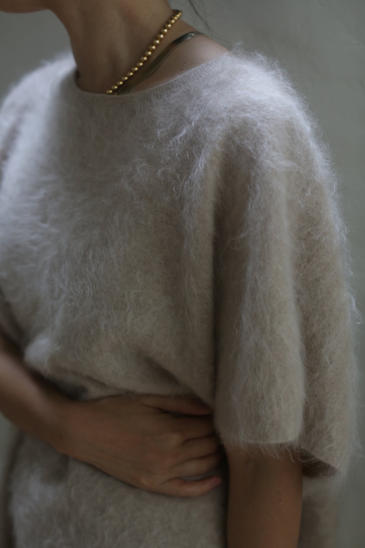 A person wearing the Salento | Sandstone, a relaxed fit, light gray sweater, and a gold chain necklace poses with one hand resting on their stomach. The background is softly lit, creating a gentle, warm atmosphere.
