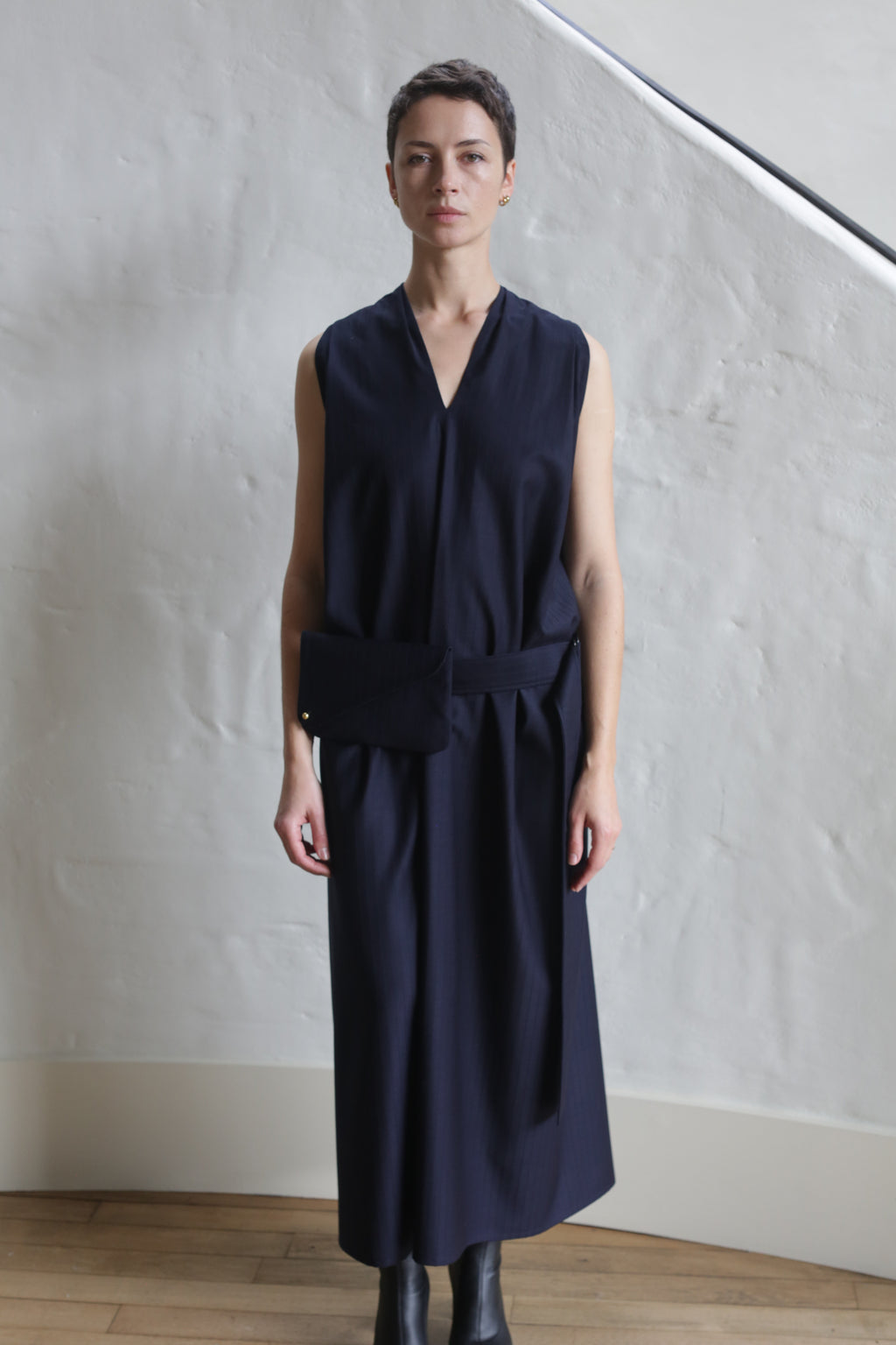 A person stands indoors against a textured white wall, wearing the Belted Dress | Wool Pinstripe Navy—a sleeveless, V-neck midi dress with a matching belt. They have short hair and are wearing black boots.
