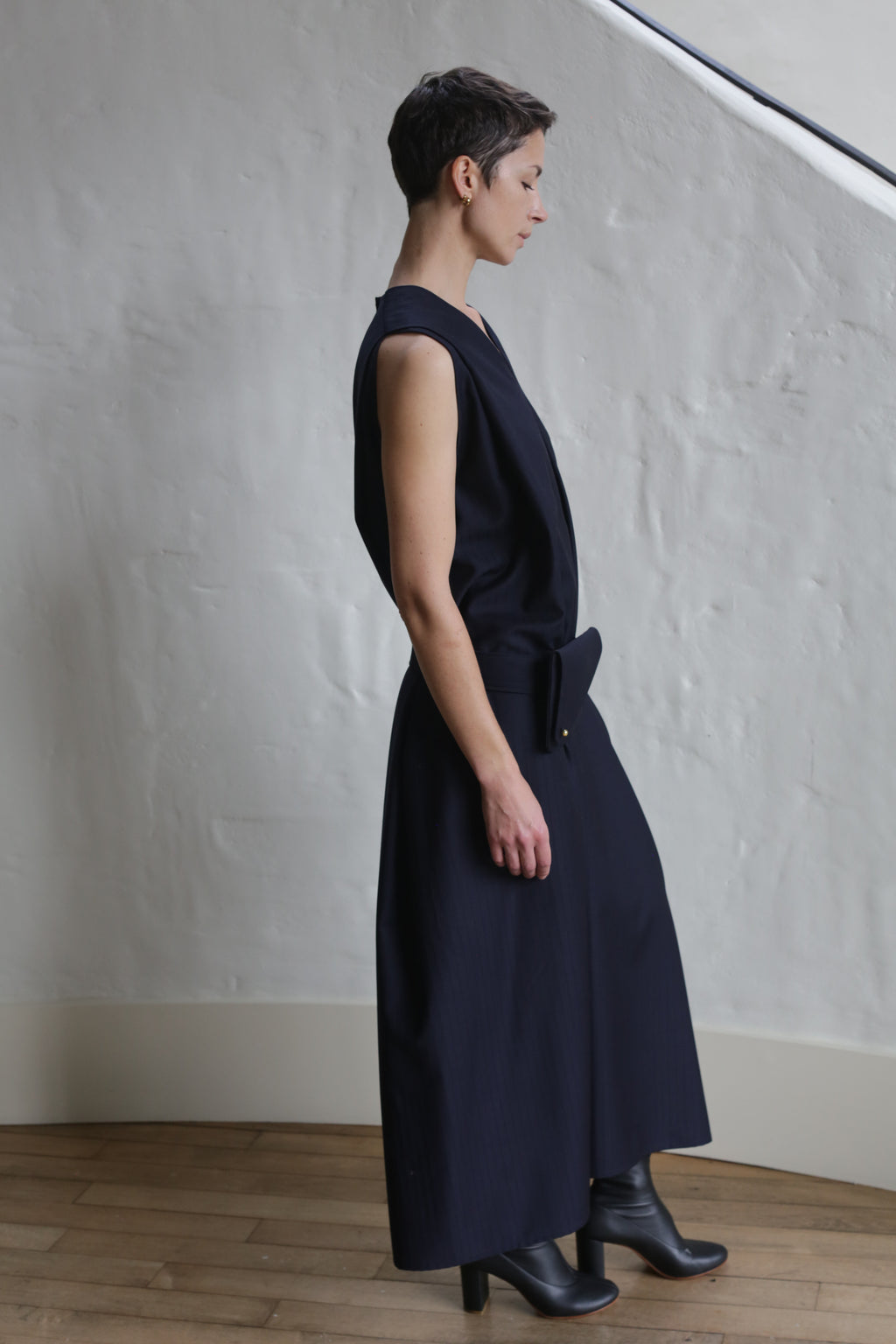 A person with short hair stands in profile against a textured white wall, wearing the Belted Dress | Wool Pinstripe Navy and black heeled boots. The scene features a wooden floor and minimal decor.