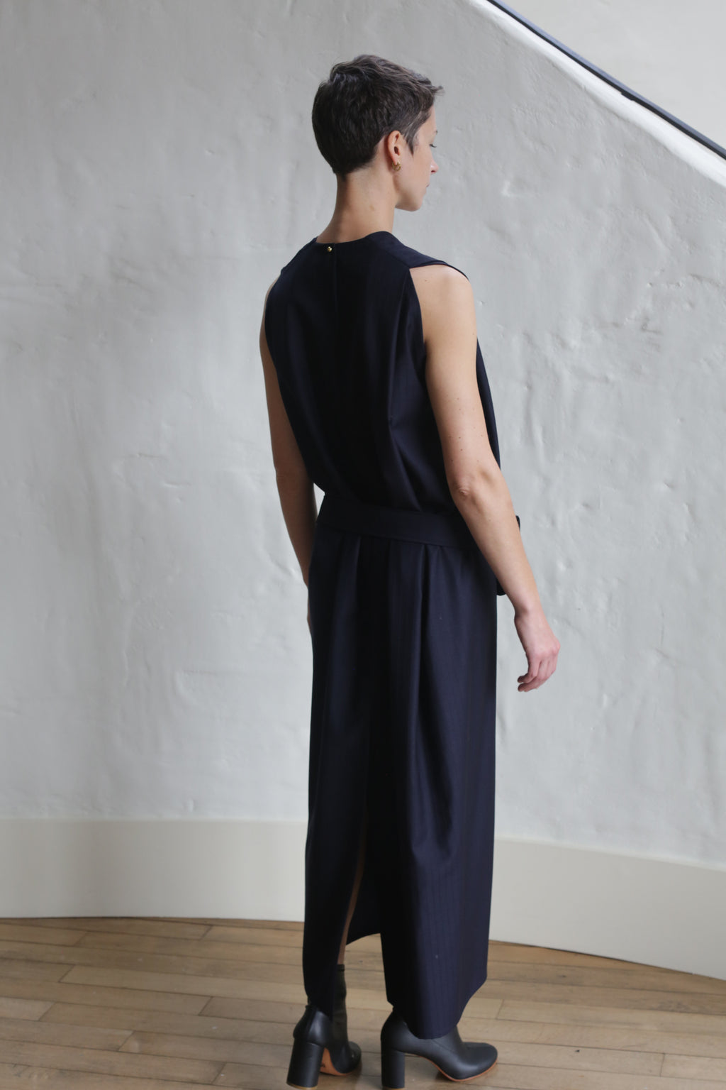 A person with short hair stands indoors facing away, wearing the Belted Dress | Wool Pinstripe Navy. The space features white textured walls, wooden flooring, and soft natural light coming from above.