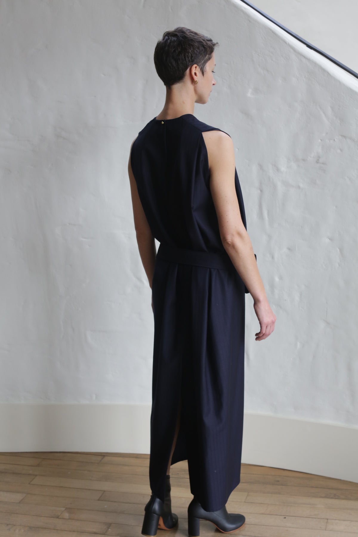 A person with short hair stands indoors facing away, wearing the Belted Dress | Wool Pinstripe Navy. The space features white textured walls, wooden flooring, and soft natural light coming from above.