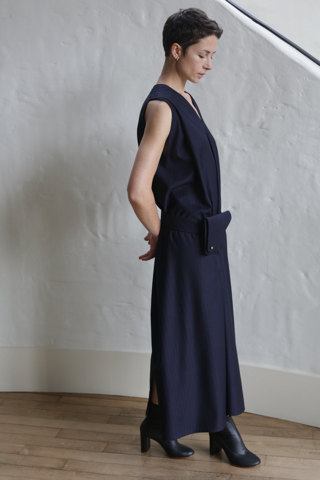 A person with short hair stands sideways in a minimalist room, wearing the Belted Dress | Wool Pinstripe Navy with a matching belt bag and black ankle boots. The background features a white textured wall and wooden floor.