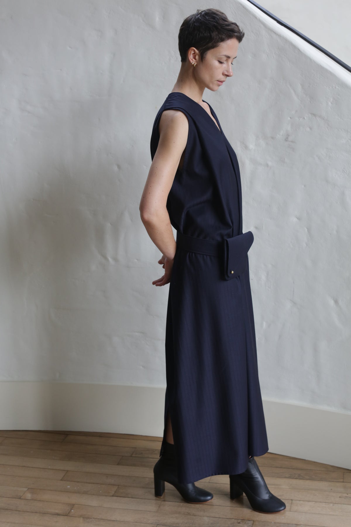 A person with short hair stands sideways in a minimalist room, wearing the Belted Dress | Wool Pinstripe Navy with a matching belt bag and black ankle boots. The background features a white textured wall and wooden floor.