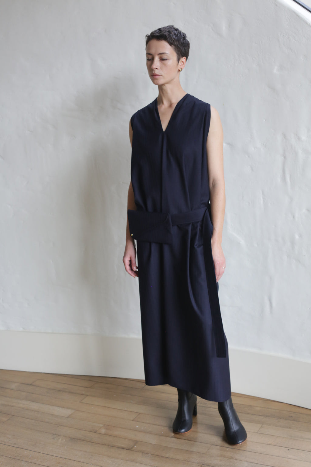 A person with short hair stands indoors against a white textured wall on a wooden floor, wearing the Belted Dress | Wool Pinstripe Navy styled with black ankle boots.