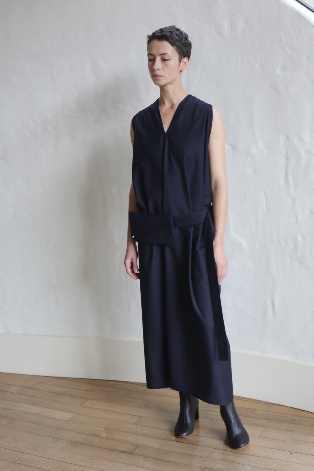A person with short hair stands indoors against a white textured wall on a wooden floor, wearing the Belted Dress | Wool Pinstripe Navy styled with black ankle boots.