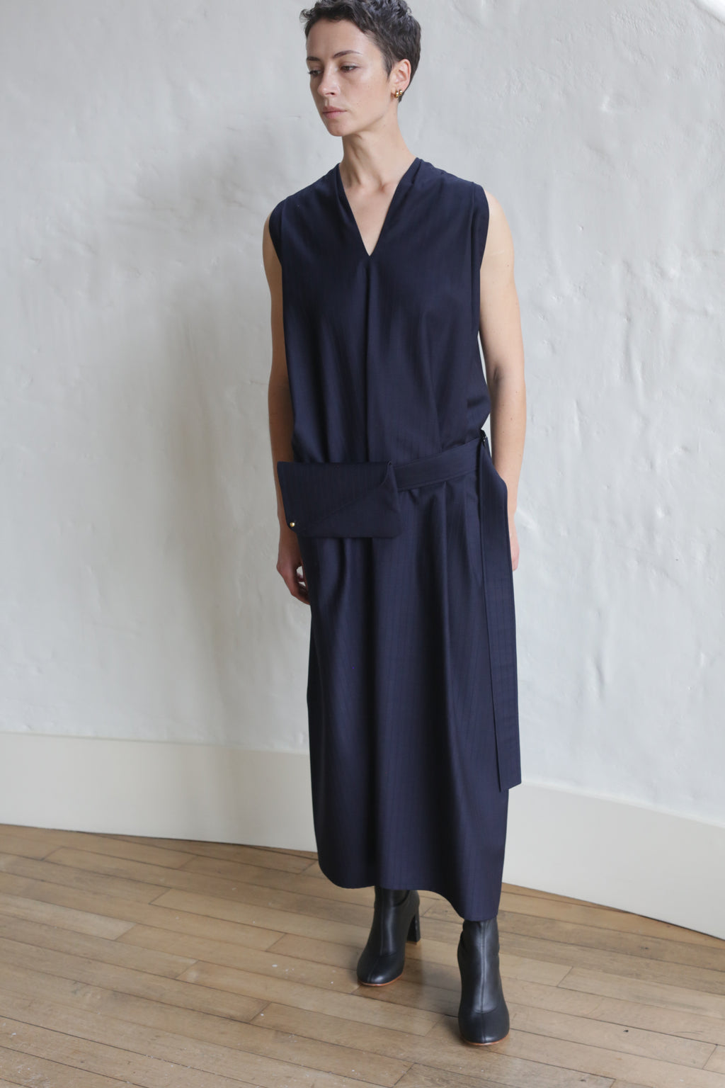 Person with short hair wearing the Belted Dress | Wool Pinstripe Navy and black ankle boots, standing on a wooden floor against a textured white wall.