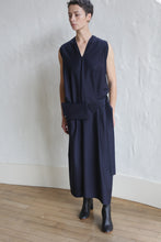 Load image into Gallery viewer, Person with short hair wearing the Belted Dress | Wool Pinstripe Navy and black ankle boots, standing on a wooden floor against a textured white wall.