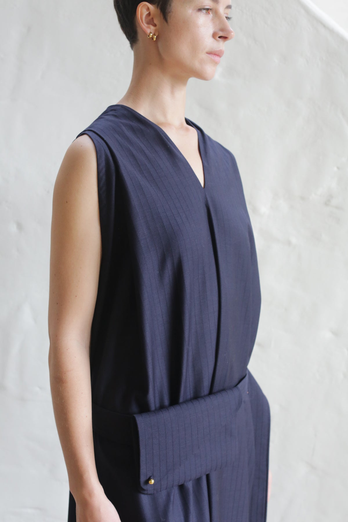 A person with short hair wears the Belted Dress | Wool Pinstripe Navy, a sleeveless dark dress with a deep V-neck and wide fabric belt, standing against a light textured wall.