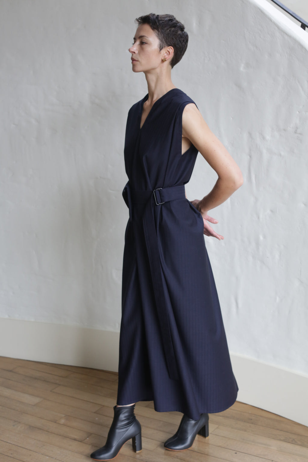 A person with short hair stands sideways against a textured white wall, wearing the Belted Dress | Wool Pinstripe Navy and black ankle boots, with their hands clasped behind their back.