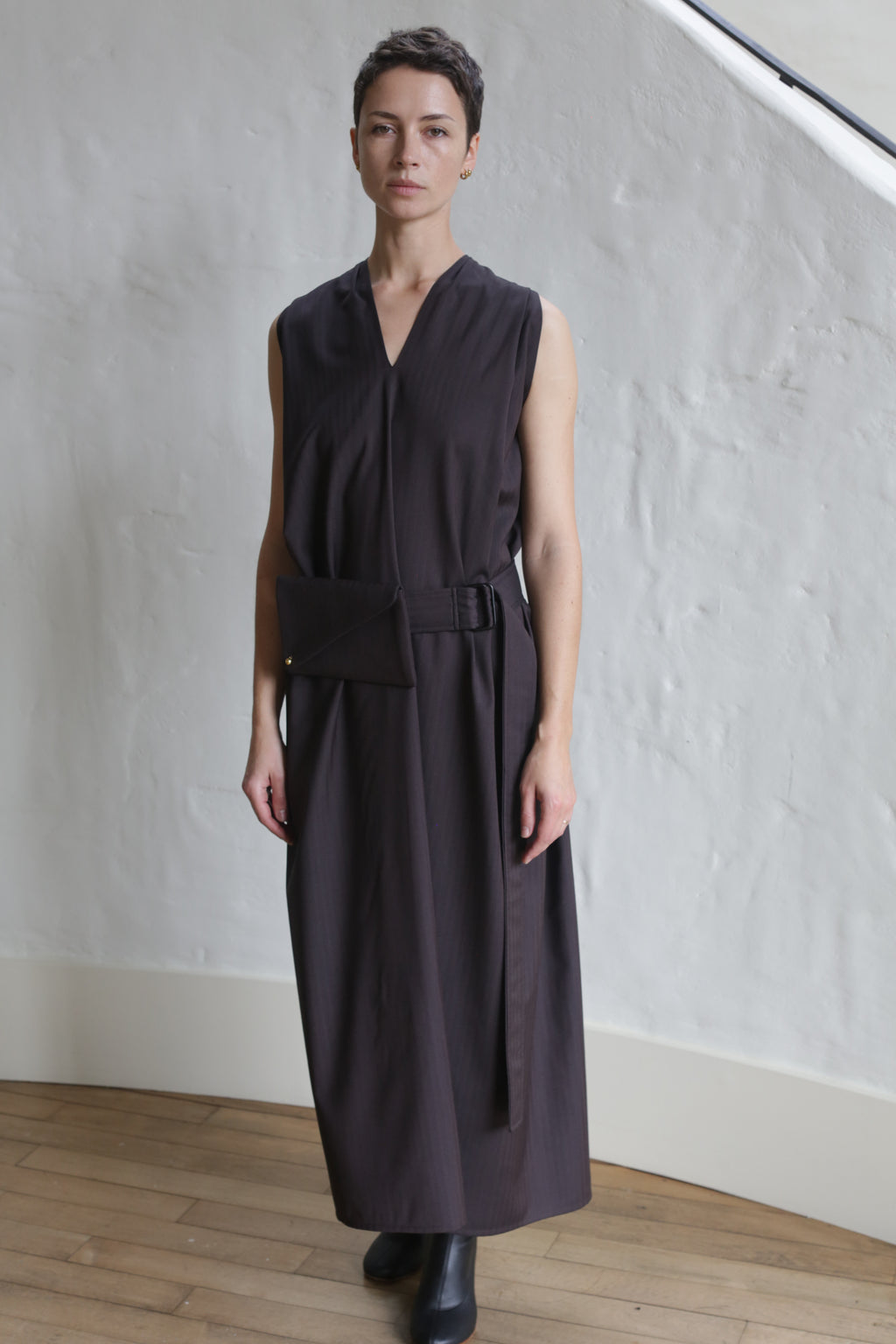 A person with short hair wears the Belted Dress | Wool Herringbone Brown, a sleeveless, ankle-length outfit with a belt and matching pouch, standing indoors on a wooden floor against a white textured wall.