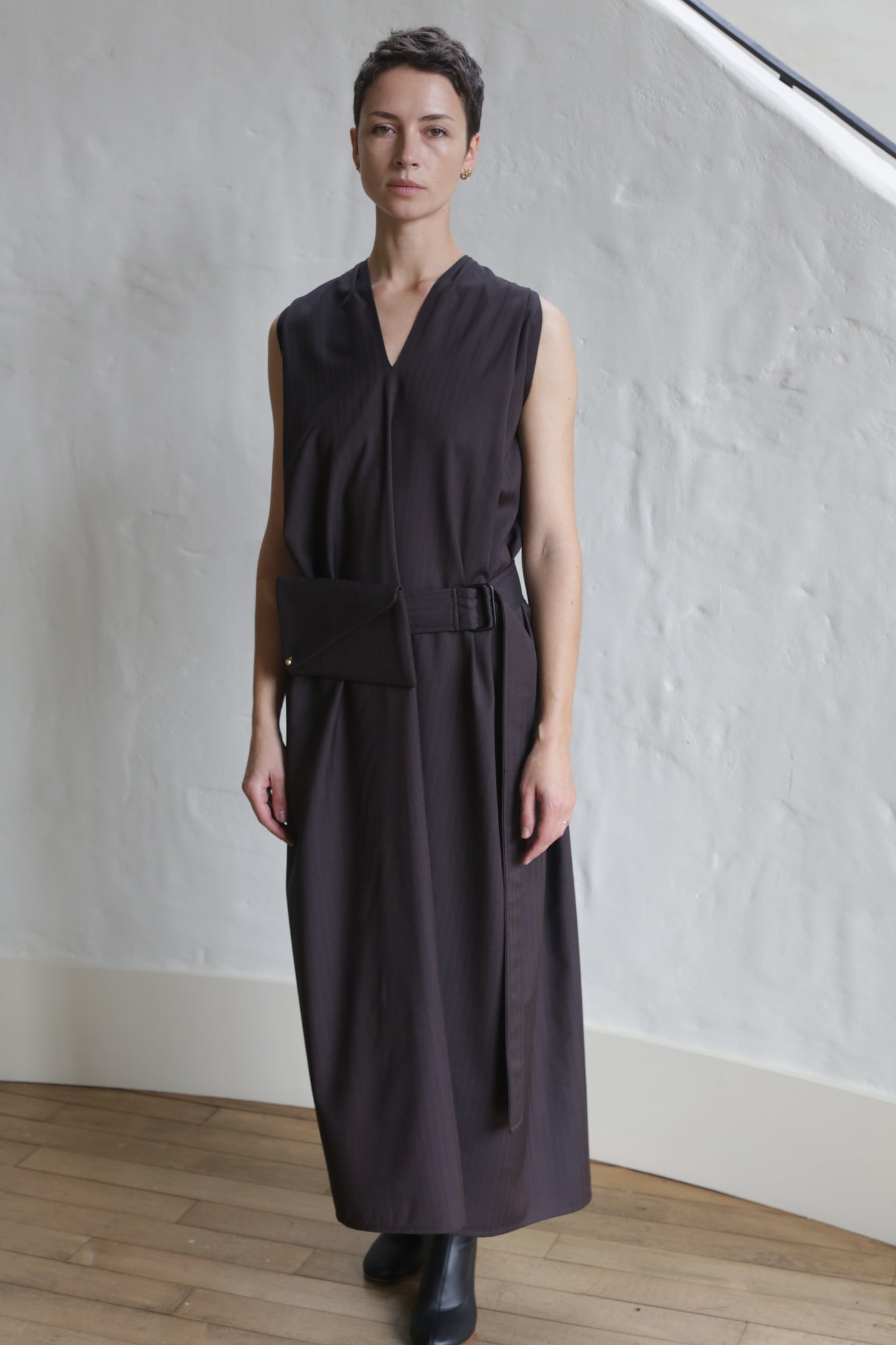 A person with short hair wears the Belted Dress | Wool Herringbone Brown, a sleeveless, ankle-length outfit with a belt and matching pouch, standing indoors on a wooden floor against a white textured wall.