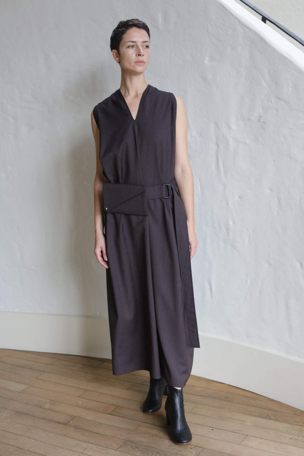 A person with short dark hair wears the Belted Dress | Wool Herringbone Brown, featuring a sleeveless, loose fit with a wide belt and rectangular pouch. They stand indoors on a wooden floor against a textured white wall.