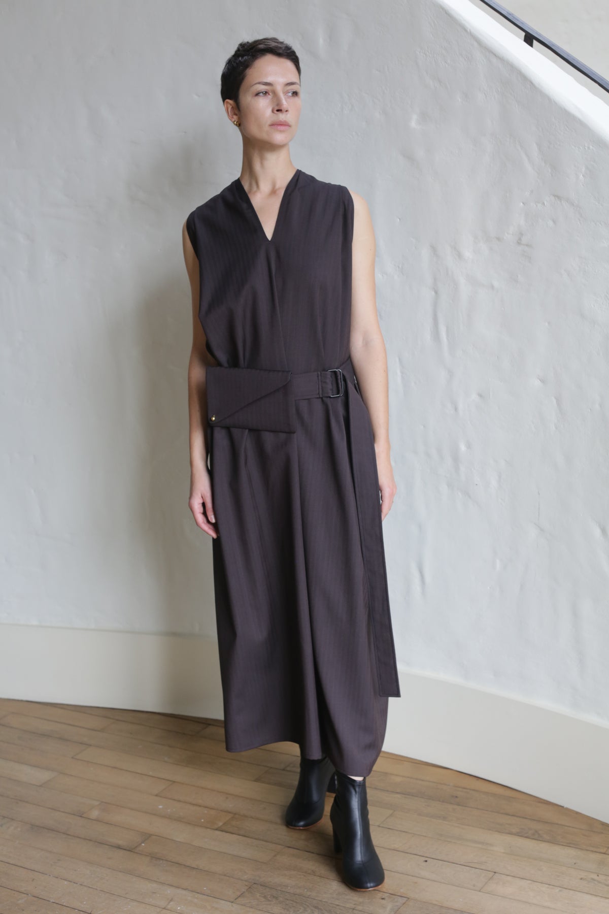 A person with short dark hair wears the Belted Dress | Wool Herringbone Brown, featuring a sleeveless, loose fit with a wide belt and rectangular pouch. They stand indoors on a wooden floor against a textured white wall.