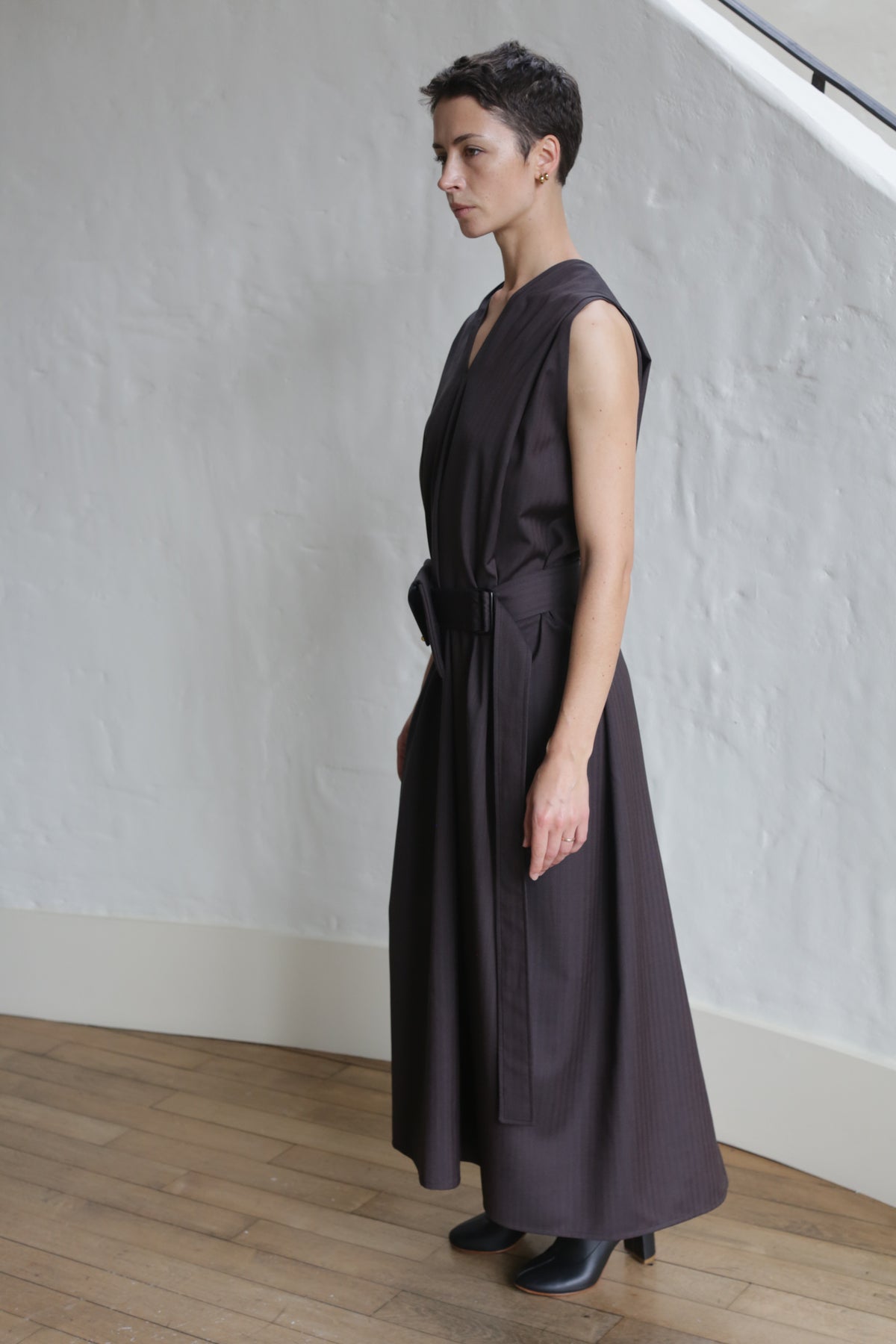 A person with short dark hair stands indoors, wearing the Belted Dress | Wool Herringbone Brown—an ankle-length, sleeveless dress with a belt—paired with black ankle boots against a white textured wall and light wood floor.