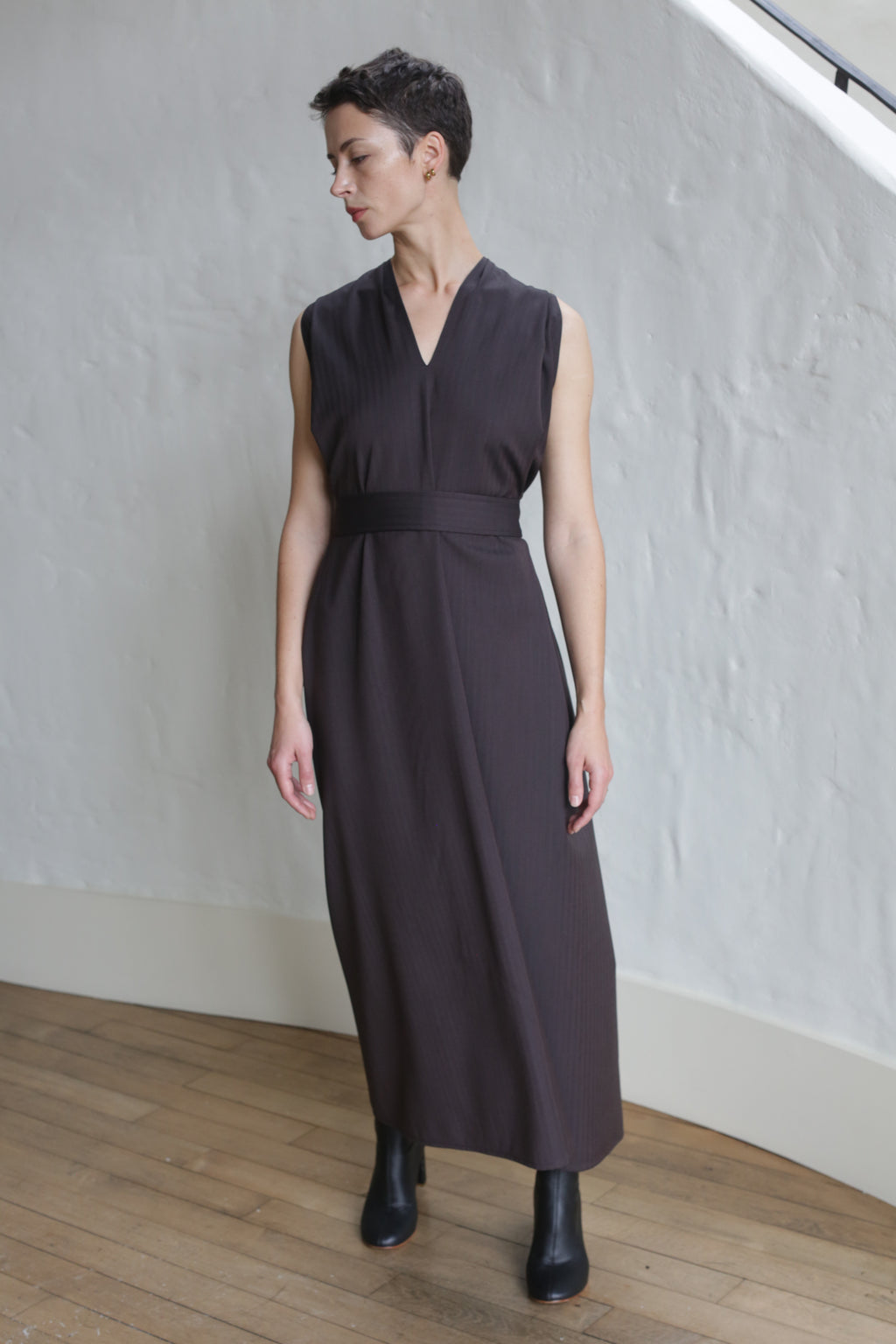 A person wearing the Belted Dress | Wool Herringbone Brown stands indoors on a wooden floor against a textured white wall, paired with black boots.