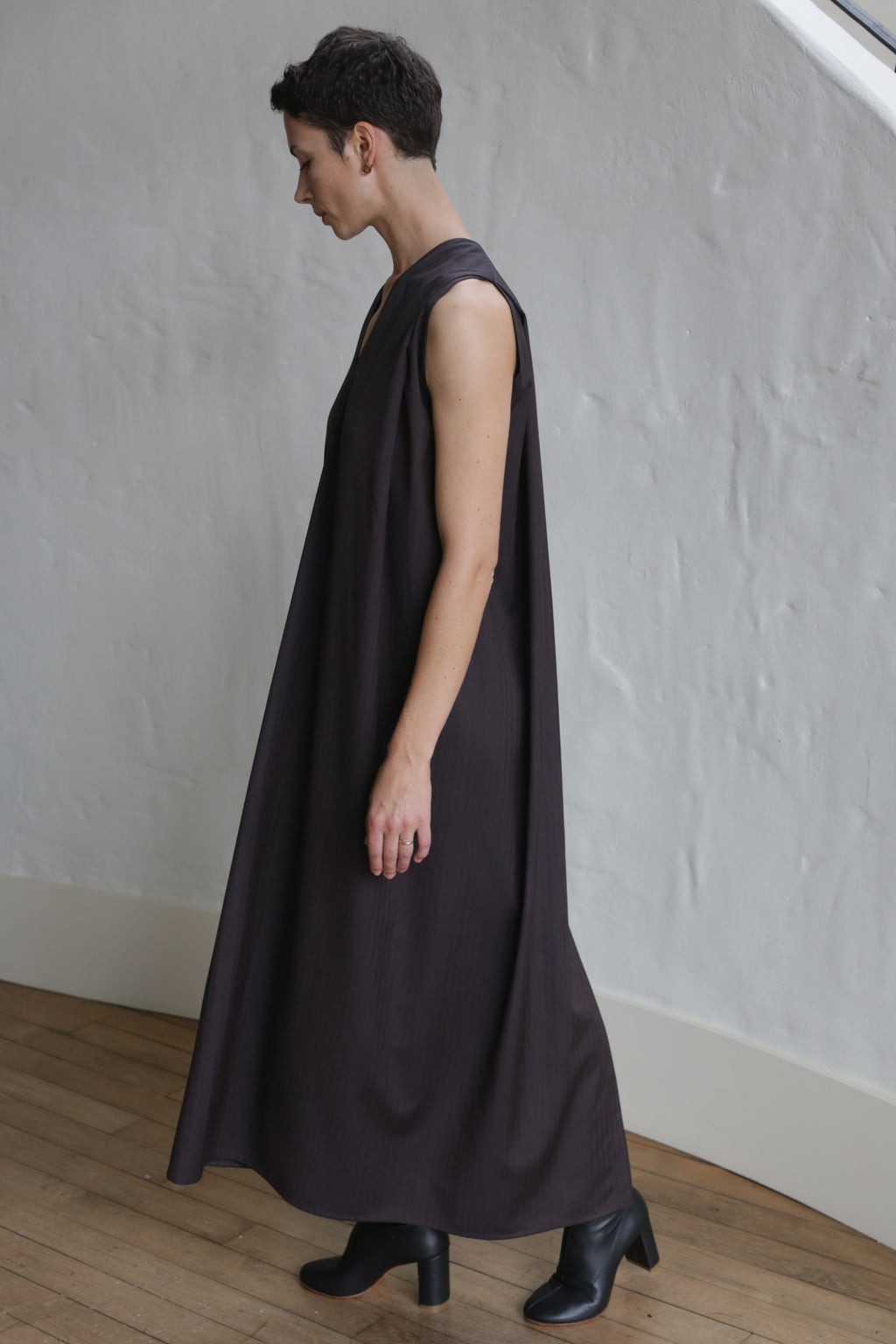 A person with short dark hair stands indoors on a wooden floor near a textured white wall, wearing the Belted Dress | Wool Herringbone Brown and black ankle boots.