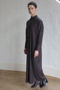 A person with short hair wears the Atelier Coat in wool herringbone brown, styled with black ankle boots, standing indoors on a wooden floor against a white textured wall.