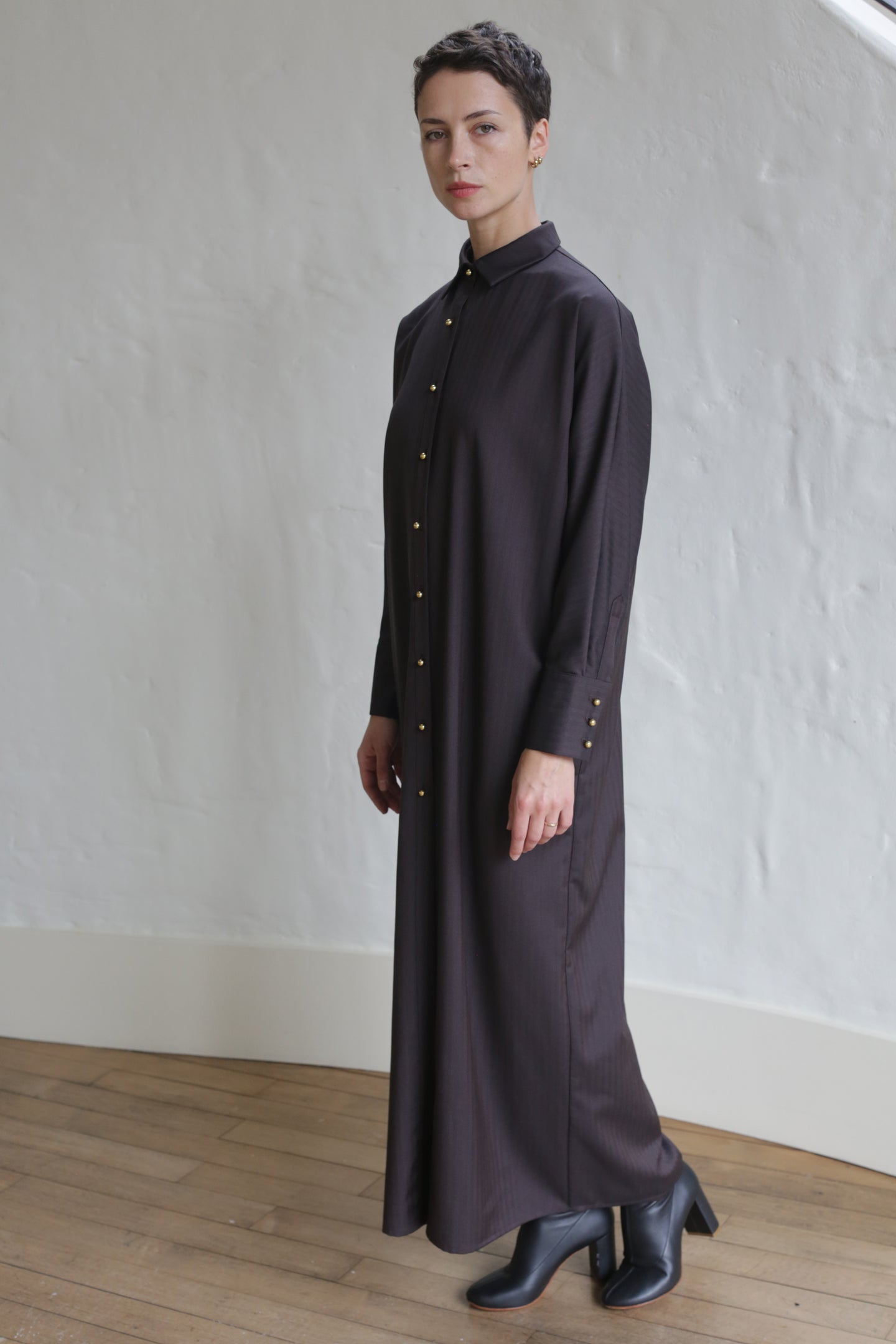 A person with short hair wears the Atelier Coat in wool herringbone brown, styled with black ankle boots, standing indoors on a wooden floor against a white textured wall.