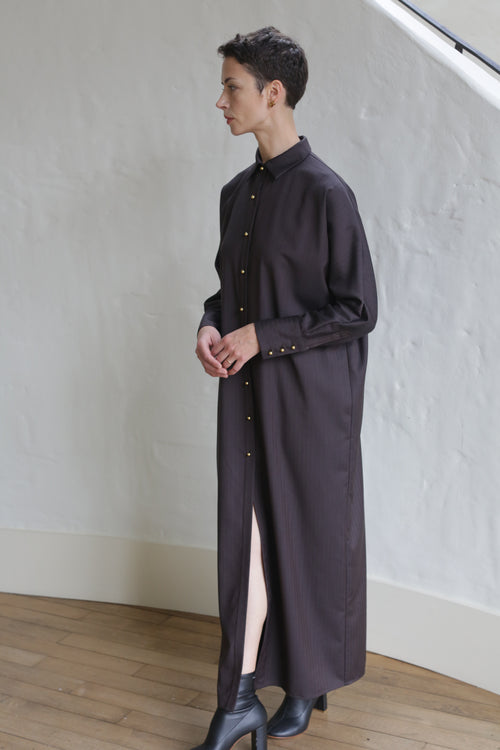 A person with short hair poses indoors in profile against a white textured wall, wearing the Atelier Coat | Wool Herringbone Brown styled as a long buttoned shirt dress with black heeled boots.