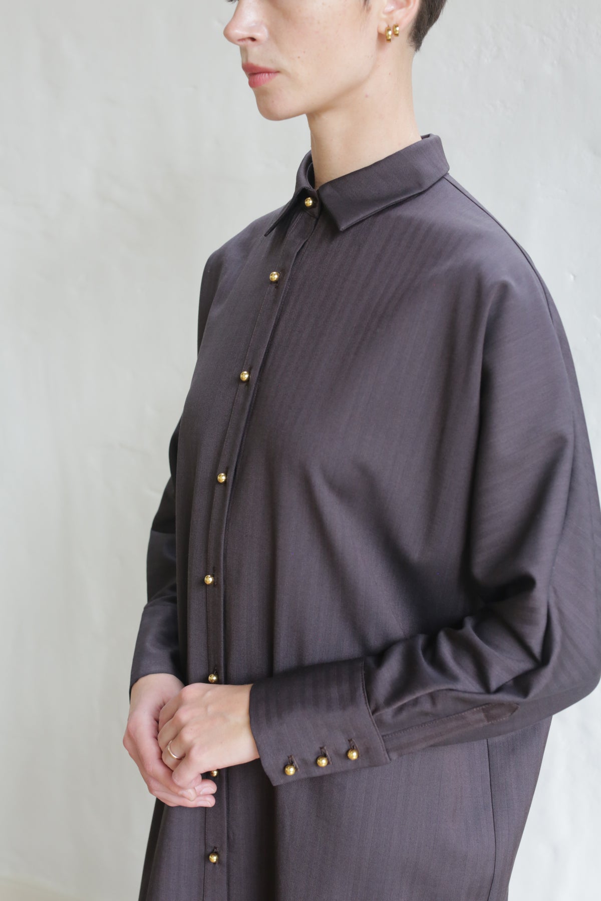 A person wearing the Atelier Coat in wool herringbone brown, a loose-fitting long-sleeved coat with gold buttons, stands against a light background. Their face is partially visible and hands are lightly clasped.
