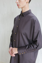 Load image into Gallery viewer, A person wearing the Atelier Coat in wool herringbone brown, a loose-fitting long-sleeved coat with gold buttons, stands against a light background. Their face is partially visible and hands are lightly clasped.