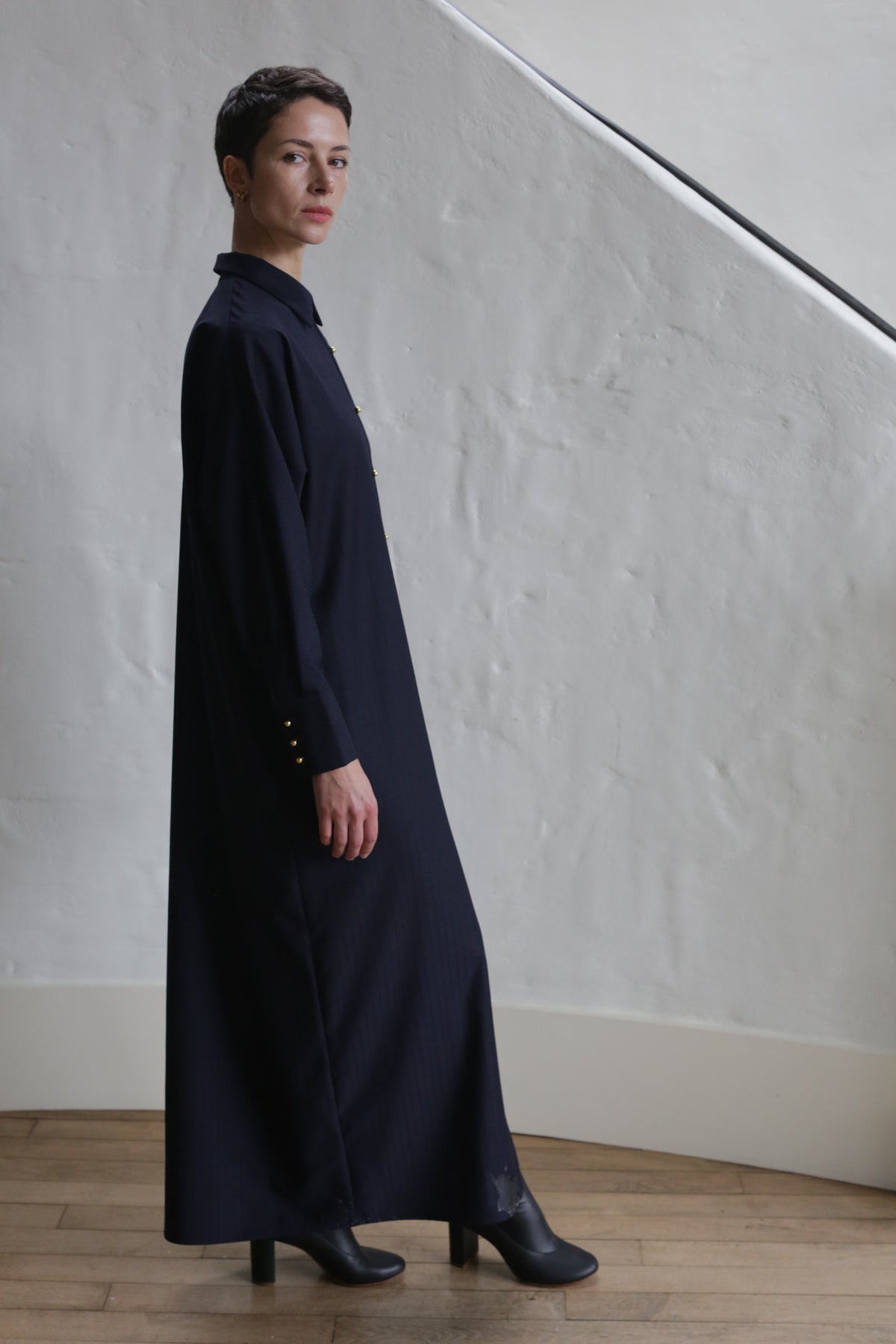 A person with short hair stands indoors on a wooden floor in front of a textured white wall and stair rail, wearing the Atelier Coat | Wool Pinstripe Navy and black boots.