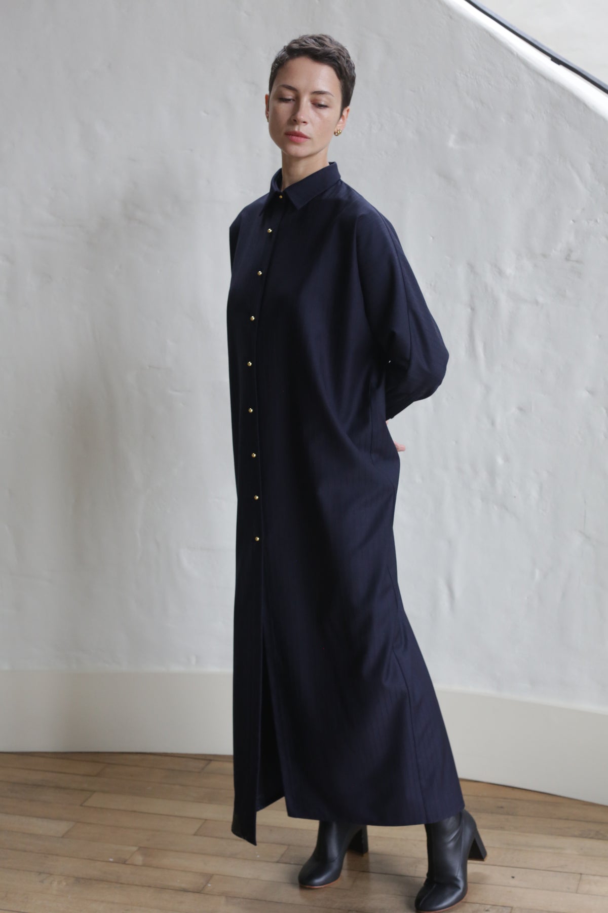 Someone stands indoors against a white textured wall and wooden floor, eyes closed and hands behind their back, wearing the Atelier Coat | Wool Pinstripe Navy with black ankle boots.