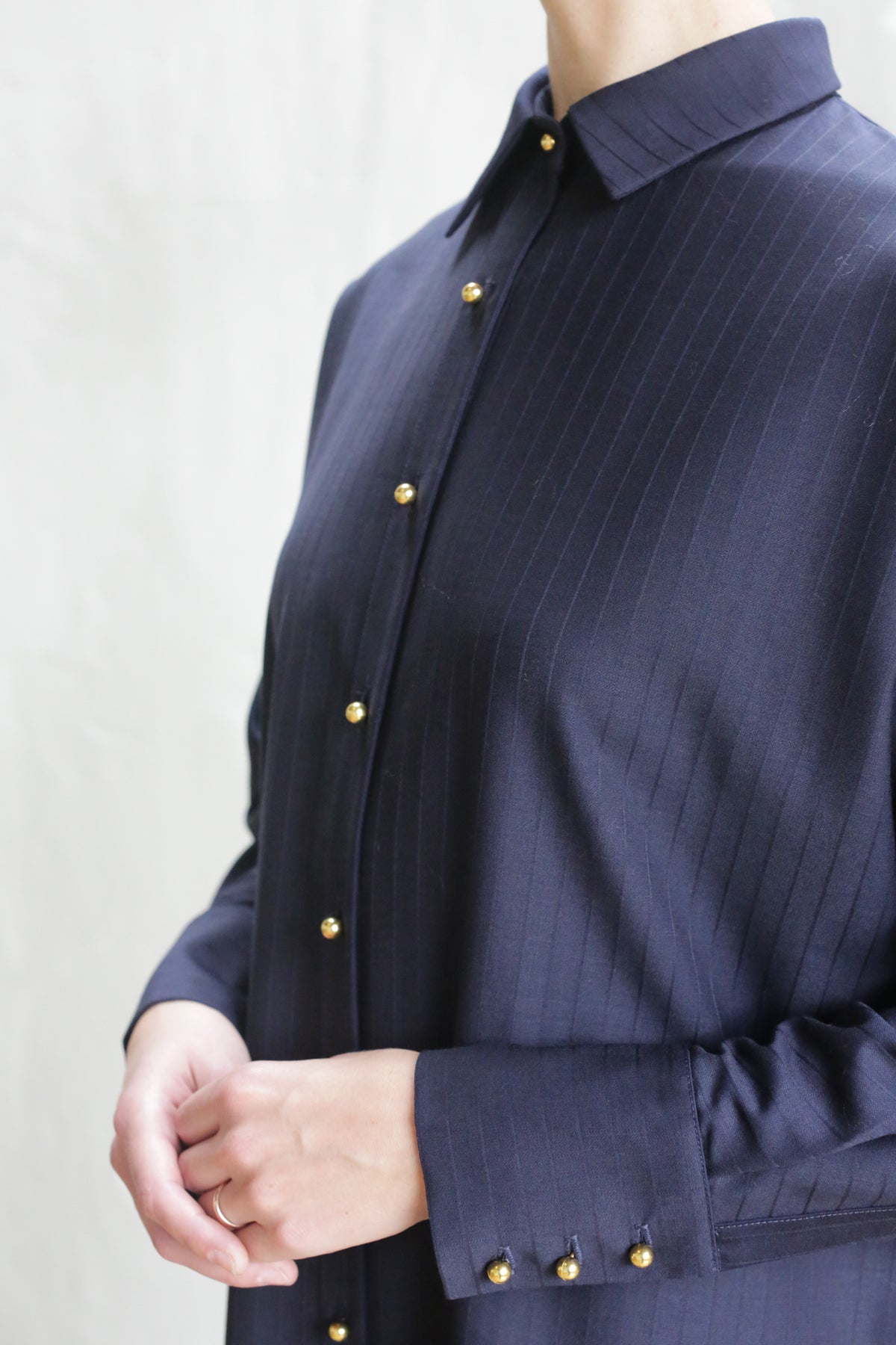 A person showcases the Atelier Coat | Wool Pinstripe Navy, featuring gold buttons, against a light background. The image highlights the coat and upper torso, with their hands gently clasped.
