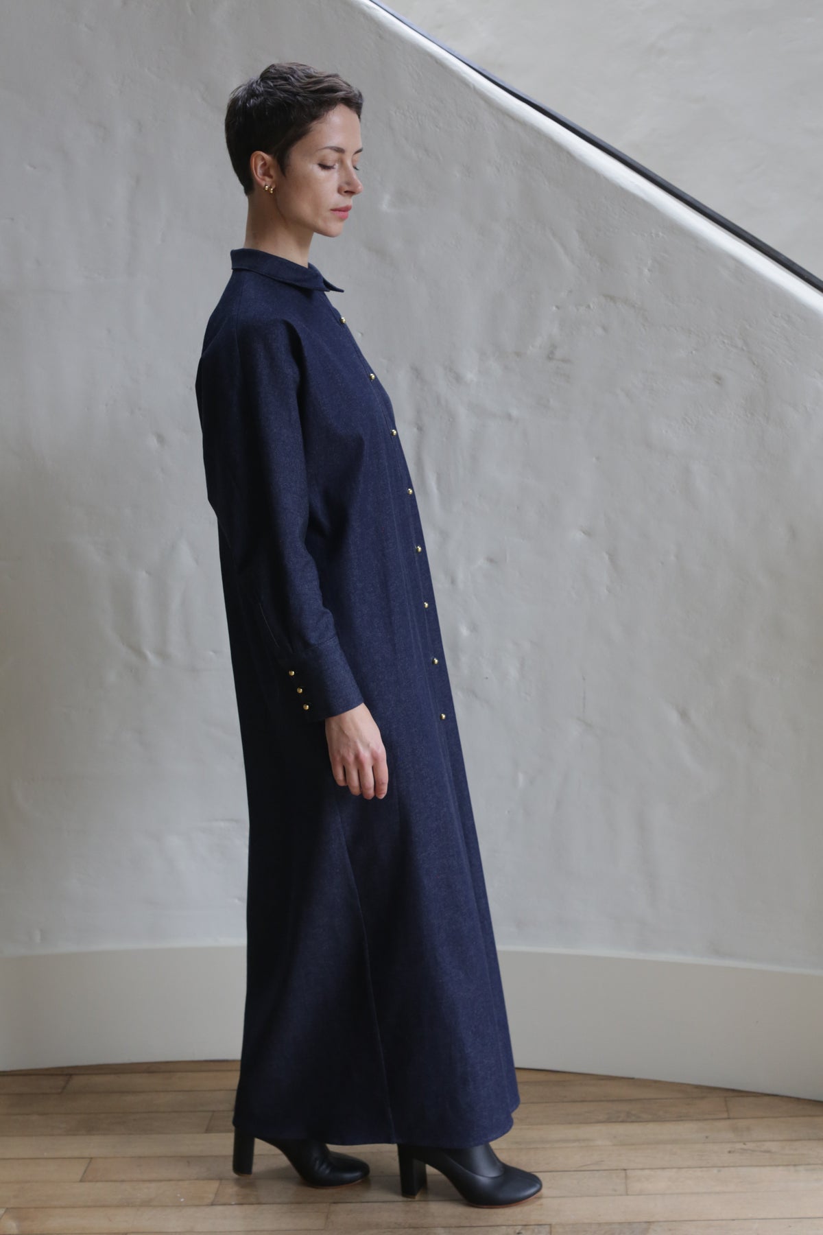 A person with short hair stands indoors facing left against a light wall, wearing the Atelier Coat | Denim Stretch—a long, dark navy button-up dress with long sleeves—paired with black heeled boots.