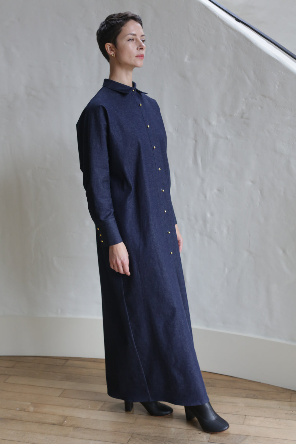 A person stands indoors wearing the Atelier Coat | Denim Stretch, a long dark blue button-down dress with long sleeves and black shoes, posing against a textured white wall and wooden floor.
