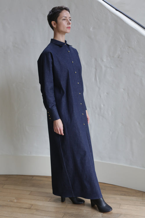 A person stands indoors wearing the Atelier Coat | Denim Stretch, a long dark blue button-down dress with long sleeves and black shoes, posing against a textured white wall and wooden floor.