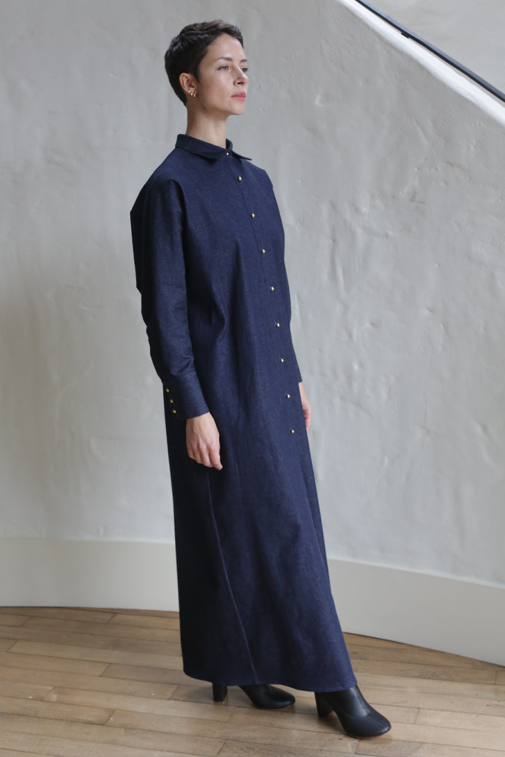 A person stands indoors wearing the Atelier Coat | Denim Stretch, a long dark blue button-down dress with long sleeves and black shoes, posing against a textured white wall and wooden floor.