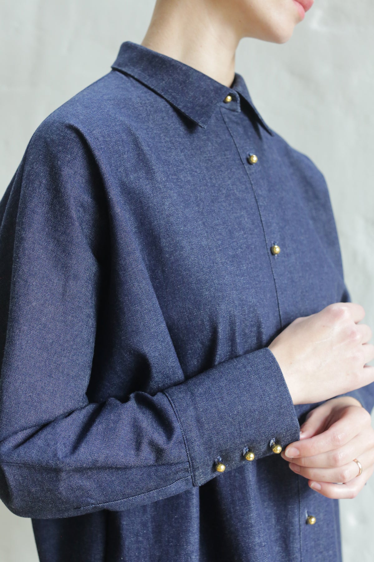 A person wears the Atelier Coat | Denim Stretch, a blue button-up featuring gold buttons. The image highlights the upper half, focusing on the collar, sleeve, and distinctive button details against a light background.