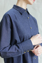 Load image into Gallery viewer, A person wears the Atelier Coat | Denim Stretch, a blue button-up featuring gold buttons. The image highlights the upper half, focusing on the collar, sleeve, and distinctive button details against a light background.