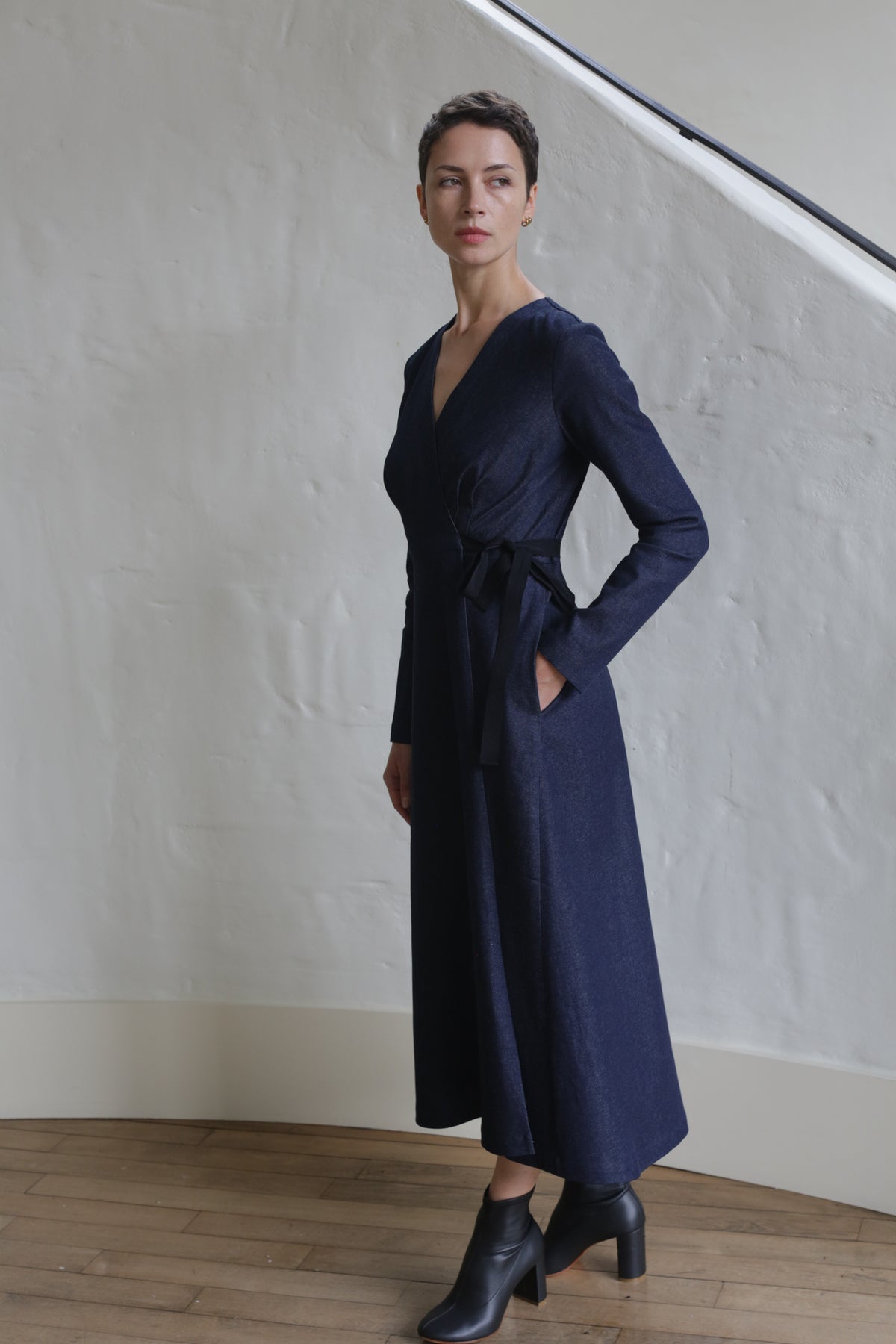 A woman with short hair stands indoors against a light textured wall, wearing the Fitted Wrap Dress | Denim Stretch in navy blue with black ankle boots, one hand in her pocket, looking slightly to the side.