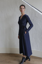 Load image into Gallery viewer, A woman with short hair stands indoors against a light textured wall, wearing the Fitted Wrap Dress | Denim Stretch in navy blue with black ankle boots, one hand in her pocket, looking slightly to the side.