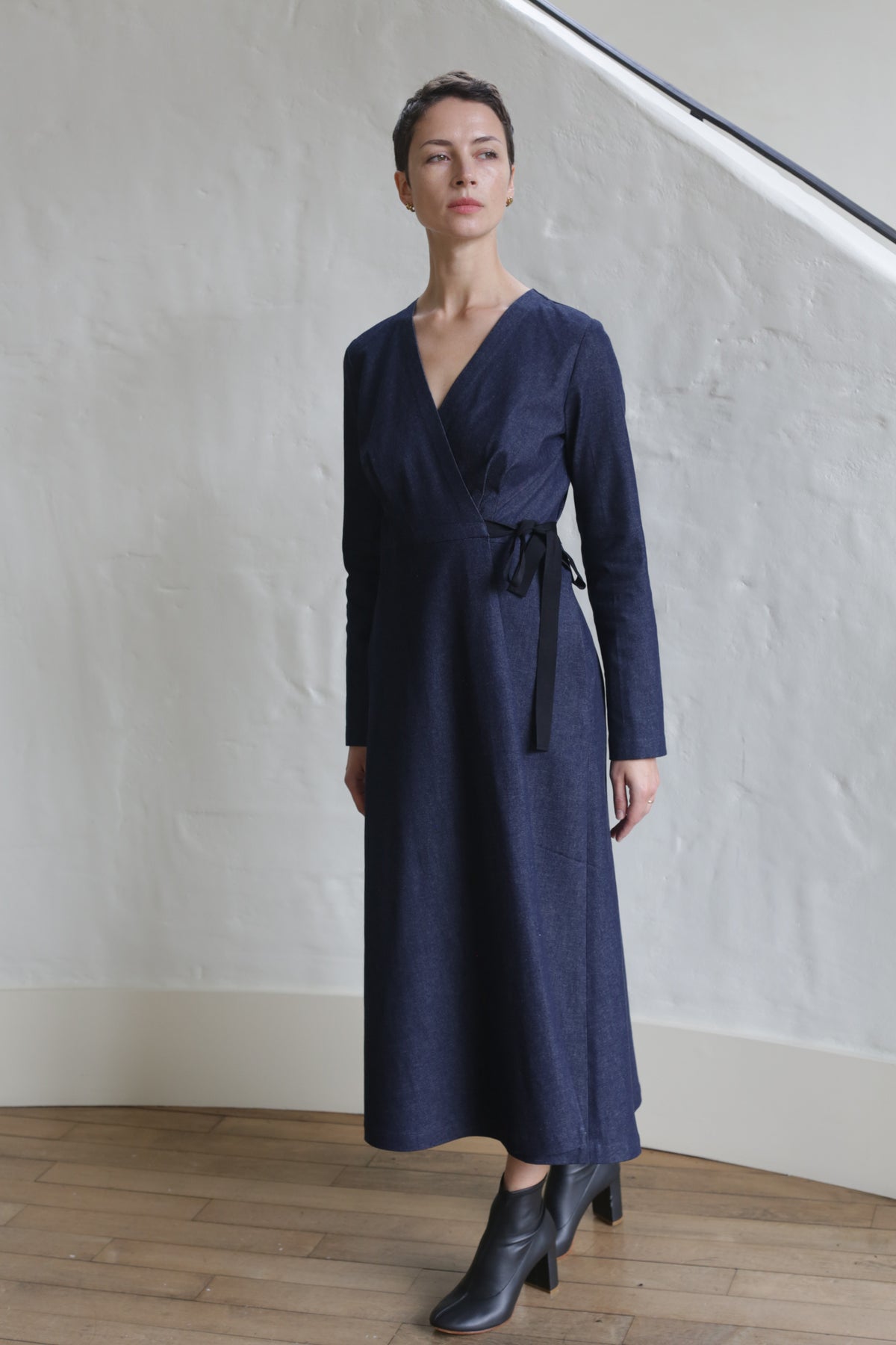 A woman stands indoors on a wooden floor against a white textured wall, wearing the Fitted Wrap Dress | Denim Stretch—a long-sleeved, navy blue wrap dress with waist tie—and black heeled ankle boots.