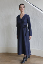 Load image into Gallery viewer, A woman stands indoors on a wooden floor against a white textured wall, wearing the Fitted Wrap Dress | Denim Stretch—a long-sleeved, navy blue wrap dress with waist tie—and black heeled ankle boots.