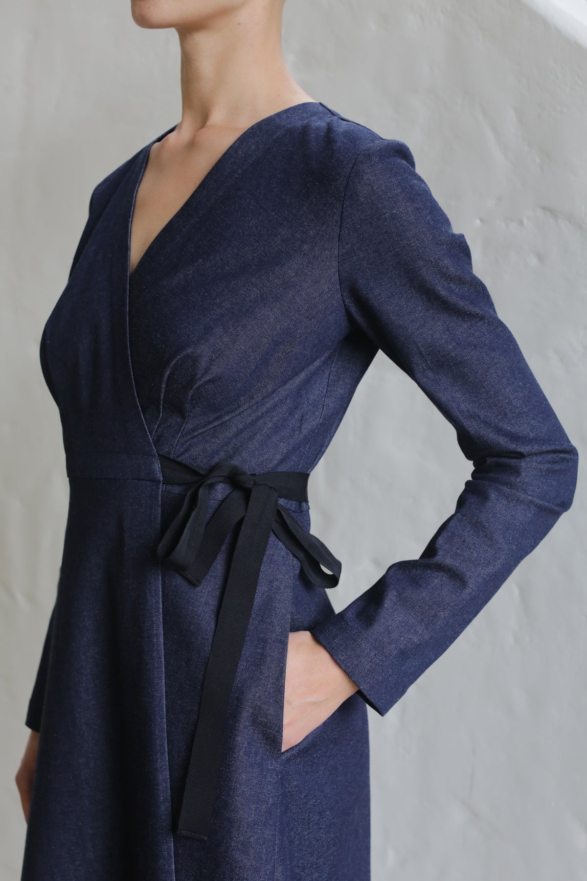 A person wearing the Fitted Wrap Dress | Denim Stretch—a long-sleeved, deep navy V-neck wrap dress with a black waist ribbon and pockets—stands against a light textured wall, hand in pocket.