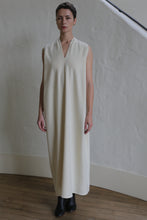 Load image into Gallery viewer, A person stands indoors in The Winter Dress | Cashmere Undyed—a sleeveless, long, cream-colored dress with a subtle V-neck—paired with black ankle boots against a light textured wall and wooden floor.