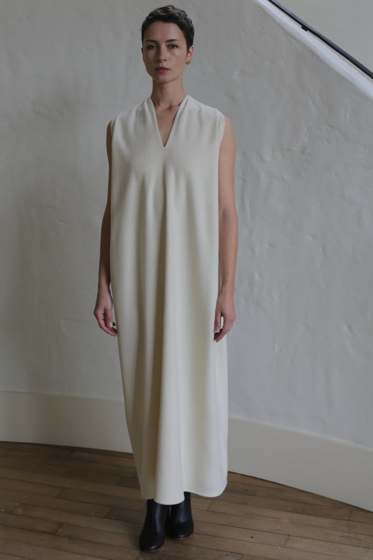 A person stands indoors in The Winter Dress | Cashmere Undyed—a sleeveless, long, cream-colored dress with a subtle V-neck—paired with black ankle boots against a light textured wall and wooden floor.