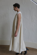 A person with short hair stands indoors, wearing The Winter Dress | Cashmere Undyed, a long, sleeveless, loose-fitting cream dress, and black shoes, facing left against a textured white wall.