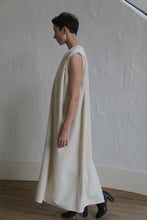 Load image into Gallery viewer, A person with short hair stands indoors, wearing The Winter Dress | Cashmere Undyed, a long, sleeveless, loose-fitting cream dress, and black shoes, facing left against a textured white wall.