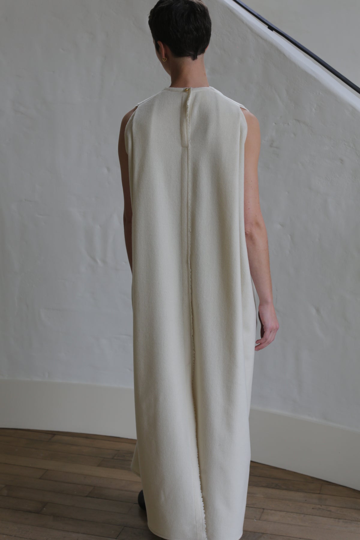 A person with short hair stands indoors, facing away, wearing The Winter Dress | Cashmere Undyed—a long, sleeveless, off-white garment with a central back seam. The setting includes a white textured wall and wooden floor.
